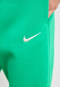 Zaļi kokvilnas sporta bikses ar baltu Nike swoosh logo, gluda tekstūra, apkaklītes potītēm un sānu kabatu labajā pusē.