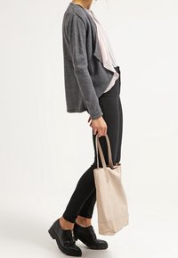 Cardigan grigio strutturato, blusa rosa chiaro, pantaloni neri slim-fit, scarpe nere con cerniera, in possesso di una borsa tote in pelle beige.