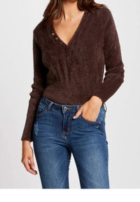 Bruine, zachte v-hals trui met drie decoratieve knopen bij de halslijn, gecombineerd met blauwe spijkerbroek met een distressed ontwerp op het dijbeen.