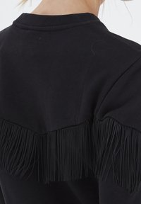 Černá mikina s třásňovým detailem podél zadních ramen, s hladkou texturou a standardním kulatým výstřihem.