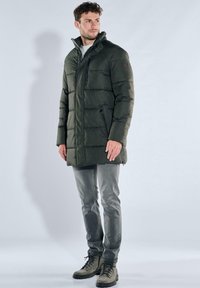 Dark green puffer coat with a high collar, zippered front pockets, and a quilted pattern. Paired with gray jeans and sturdy boots.