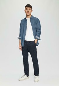 Chemise bleue clair à boutons sur un t-shirt blanc, associée à un pantalon cargo foncé et des baskets blanches. Tissu lisse avec une coupe décontractée.