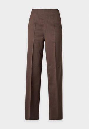 Kvinde iført brune bukser med vide ben, matchende brun blazer, pink skjorte med knapper og hvide spidse hæle står foran en ensfarvet baggrund.