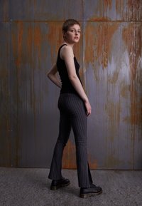 Black sleeveless top paired with gray pinstriped flared pants. Model wears black lace-up boots. Background features textured metallic wall.