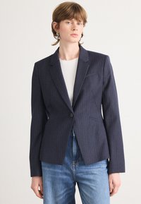 Navy modrý pruhovaný blazer s přiléhavým střihem, zapínáním na jeden knoflík a klopovým límečkem. Kombinováno se světle modrými džíny.