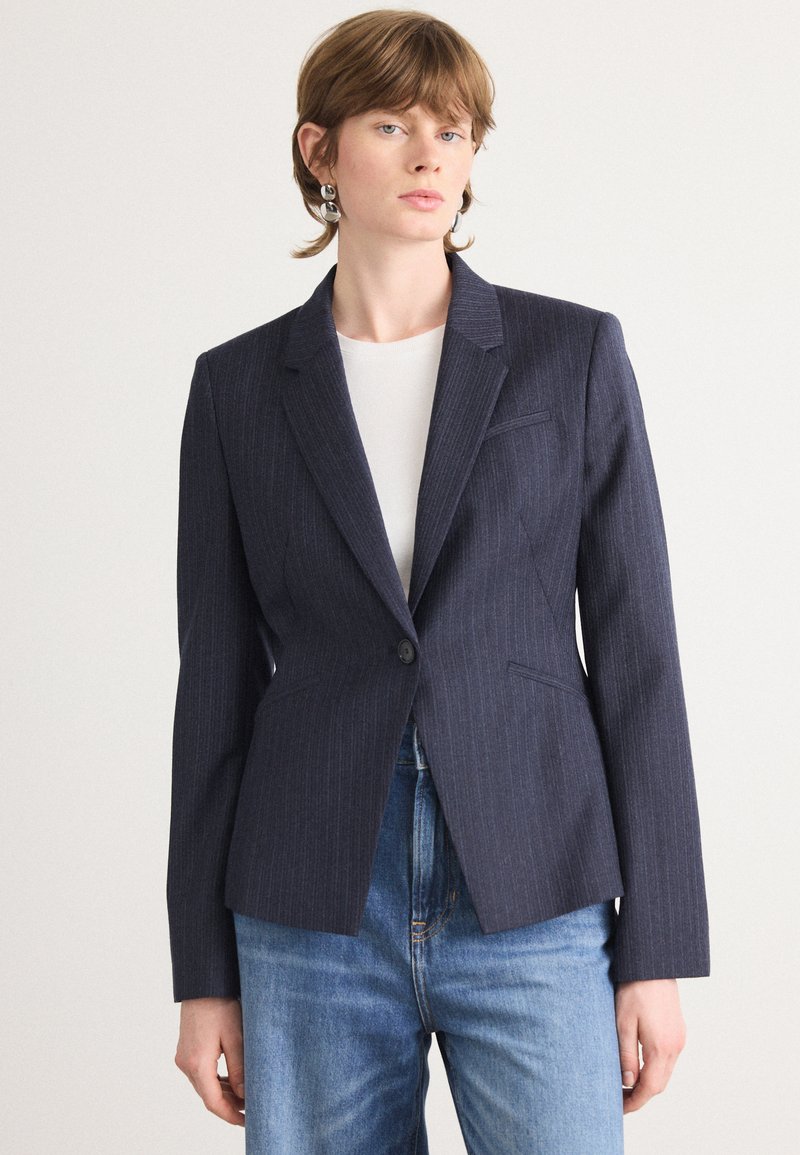 Navy modrý pruhovaný blazer s přiléhavým střihem, zapínáním na jeden knoflík a klopovým límečkem. Kombinováno se světle modrými džíny.
