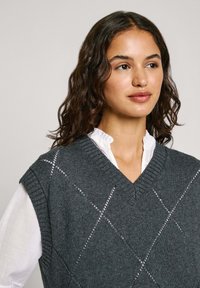 Gilet grigio a maglia con scollatura a V e motivo a rombi, indossato sopra una camicia bianca con colletto. Maniche corte e dettagli a coste sul collo.