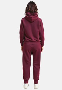Bordowa bluza z kapturem i zestaw joggerów, wykonane z miękkiego materiału, z prążkowanymi mankietami i luźnym krojem. Białe sneakersy dopełniają całość.