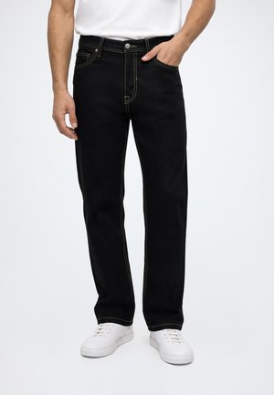 Homme portant un jean droit noir avec des coutures contrastantes, des baskets blanches et une chemise blanche, debout avec une main dans la poche.