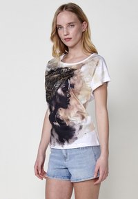 T-shirt en coton blanc avec un motif abstrait noir et marron représentant un profil avec un couvre-chef géométrique. Manches courtes et col rond.