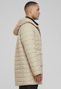 Beige quiltet dunjakke med hætte, der har en frontlynlås og lomme detaljer, vist fra siden på en neutral baggrund.