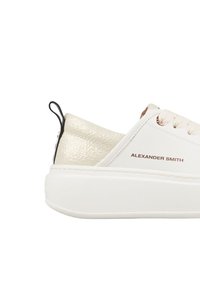 Sneaker bianco con accento dorato testurizzato, suola piatta e lacci beige. Marchio "ALEXANDER SMITH" in marrone vicino alla cucitura laterale.