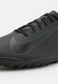Black athletic shoe with a textured upper, featuring horizontal grooves and a teal Nike swoosh. Outsole has a rugged pattern for traction.