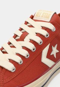 Sneaker in tela rossa con lacci beige, occhielli argentati e suola in gomma beige. Presenta un logo a stella bianca e l'etichetta "ALL STAR" sulla linguetta.