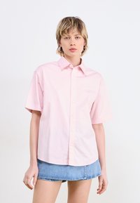Camicetta a maniche corte di colore rosa chiaro, con chiusura a bottoni, colletto a punta e dettagli ricamati discreti. Abbinata a una minigonna di jeans.