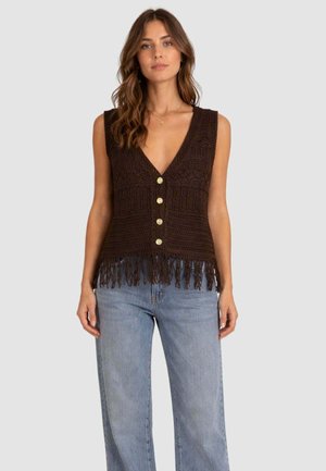 Vrouw draagt een mouwloze donkerbruine gehaakte vest met knopen en franjes aan de zoom, gecombineerd met lichtblauwe spijkerbroeken.