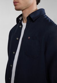 Camicia blu navy con bottoni, realizzata in tessuto testurizzato, dotata di taschino sul petto con un piccolo dettaglio a forma di bandiera e colletto a ribalta. T-shirt bianca sotto.