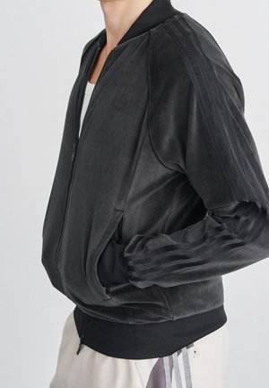 Personne portant une veste zippée en velours noire avec poignets et ourlet côtelés, avec trois bandes sur les manches et des poches latérales.