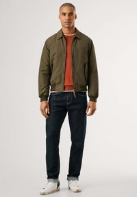 Chaqueta bomber de color verde oliva con cremallera frontal, puños de canalé, usada sobre un suéter color óxido y jeans oscuros, combinada con zapatillas blancas.
