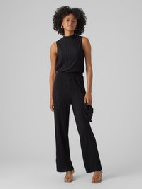 Combinaison noire plissée avec un col montant et une conception sans manches. Jambes évasées et taille cintrée. Le modèle tient un petit accessoire noir.