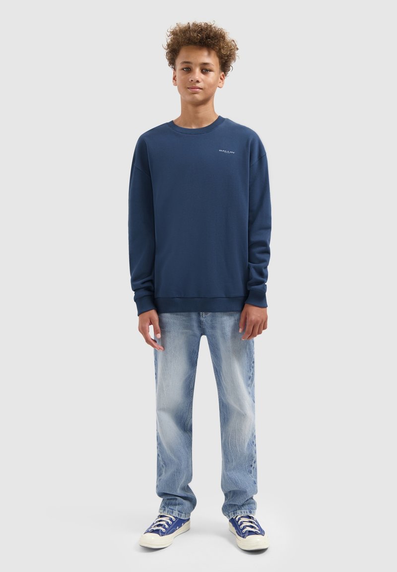 Tienerjongen staat voor een effen lichtgrijze achtergrond, gekleed in een marineblauwe sweatshirt, lichtblauwe spijkerbroek en blauwe sneakers.