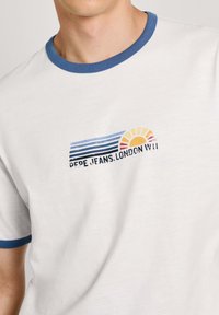 Balta marškinėliai su mėlynu apykaklės kraštu, ant kurių yra grafika su horizontaliomis juostelėmis, saule ir tekstu: "PEPE JEANS.LONDON W11."