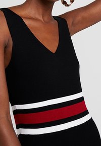 Robe en maille noire avec un décolleté en V, présentant des rayures horizontales rouges, blanches et noires à la taille. Tissu texturé avec une silhouette ajustée.