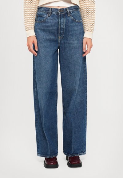 Jean bleu à jambe large en denim, avec une taille haute, des poches avant et une coupe décontractée. Tissu texturé avec des décolorations subtiles.