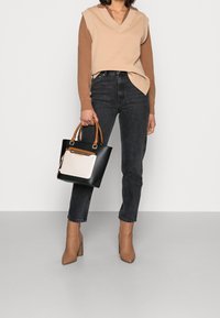 Kobieta nosząca beżowy bezrękawnik na brązowej bluzie z długim rękawem, czarne jeansy z wysokim stanem, beżowe botki na obcasie, trzymająca czarno-białą torebkę.