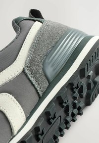 Grau-weißer Sneaker aus strukturiertem Wildleder, mit einem Kunststoff-Hackenschutz und einer schwarzen, robusten Gummisohle, schräg auf einem hellen Hintergrund platziert.
