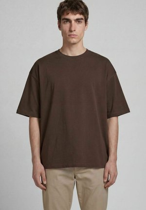 Giovane uomo che indossa una t-shirt semplice marrone scuro oversize a maniche corte e pantaloni beige chiaro, in piedi davanti a uno sfondo chiaro uniforme.