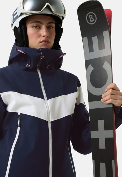 Granatowa kurtka narciarska z białym ukośnym paskiem, kieszeniami na zamek i wysokim kołnierzem. Narty trzymane w ręku z wyraźnym logo. Zestaw wyposażony w kask i gogle.