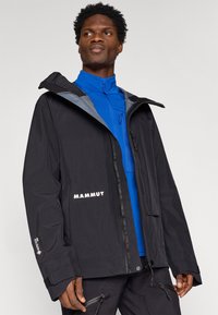 Czarna wodoodporna kurtka z zamkiem błyskawicznym z przodu, regulowanym kapturem i detalem z logo. Posiada teksturowaną niebieską wewnętrzną warstwę i widoczne elementy metalowe.