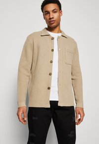 Cardigan texturé beige avec un col, fermeture à boutons, poche poitrine et manches longues. Associé à une chemise blanche et un jean noir.