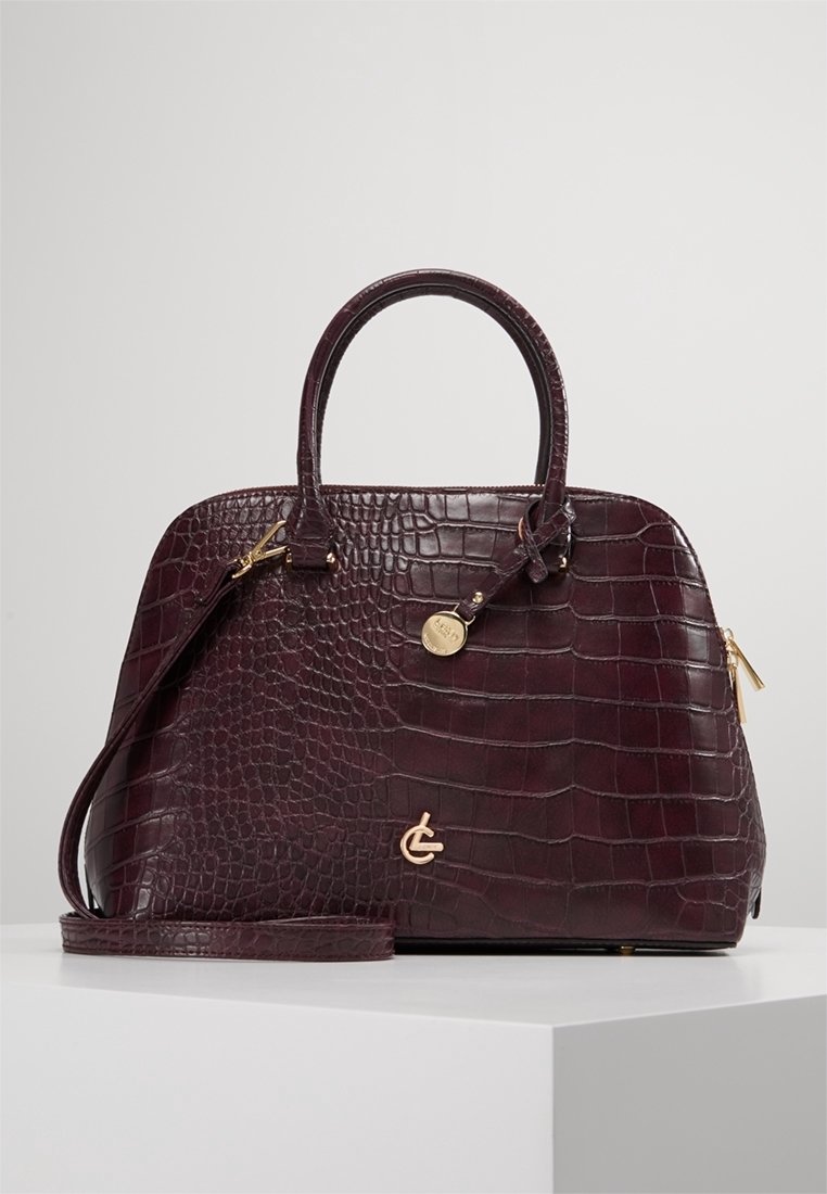 Sac à main en cuir de couleur bordeaux foncé à motif crocodile, avec des poignées supérieures, une bandoulière amovible, et des détails en métal doré sur la fermeture éclair et le logo.