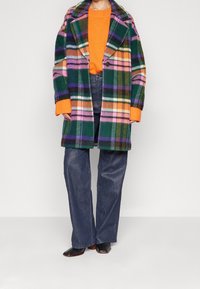 Person i en farverig overdimensioneret plaidfrakke over en orange sweater, parret med mørkeblå bredbenede jeans og sorte sko med firkantet tå.