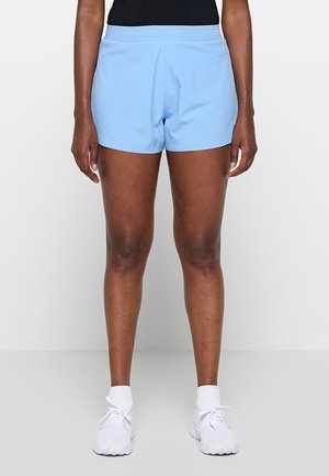 Personne portant un short de sport bleu clair, des chaussettes blanches et des chaussures de course blanches, debout devant un fond clair uni.