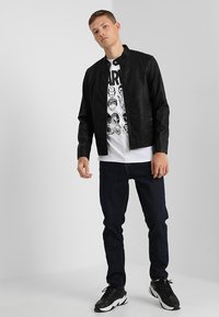 Sort faux læderjakke med trykknapkrave, iført over en hvid t-shirt med grafisk tryk med tegneseriefigurer, parret med mørkeblå jeans og sorte sneakers.