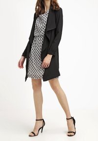 Svart kappa med öppning fram över en vit klänning med ett svart abstrakt mönster. Klänningen har en rund halsringning. Svarta högklackade sandaler.