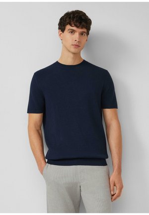 Młody mężczyzna z krótkimi, kręconymi włosami, ubrany w granatowy, krótki sweter dzianinowy z krótkim rękawem oraz jasnoszare spodnie, stojący na tle jasnej, gładkiej ściany.