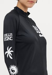 Črni rash guard z visokim ovratnikom, polnim zadrgo, belim logotipom in grafikami na rokavih; ima gladko, raztegljivo teksturo.