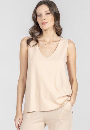 Kobieta o falowanych brązowych włosach, ubrana w bezrękawnik w kształcie litery V w kolorze beżowym oraz pasujące beżowe spodnie, pozująca z jedną ręką w kieszeni.