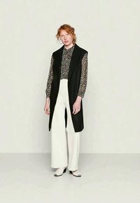 Colete preto sem mangas, camisa estampada por baixo, calças brancas largas e sapatos de salto bloco. Tecido suave, design ajustado e cores neutras.