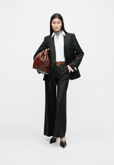 Zwarte op maat gemaakte blazer over een wit shirt en grijze coltrui, gecombineerd met wijde zwarte broeken, een bruine leren riem en een bruine tas met gouden accessoires.