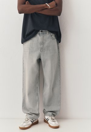 Person steht mit verschränkten Armen, trägt hellgraue lockere Jeans, schwarze Oberteil, silbernes Armband und weiße Sneaker mit schwarzen Streifen.