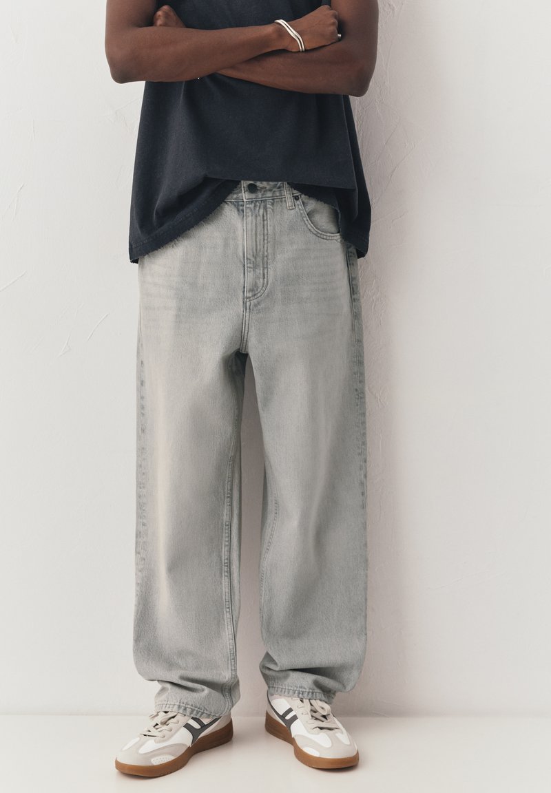 Personne debout, les bras croisés, portant un jean ample gris clair, un haut noir, un bracelet en argent et des baskets blanches avec des rayures noires.