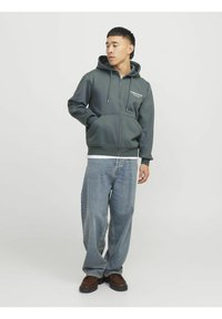 Sudadera gris con cremallera, cordones y bolsillos frontales, superpuesta a una camiseta blanca, combinada con unos jeans azules holgados y calzado marrón.