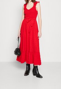 Punainen maksimekko, jossa on kuminauhahelma, frillaolkaimet ja solmittava vyö. Hameessa on kerroksellinen helma. Mustat nilkkasaappaat ja pieni laukku.