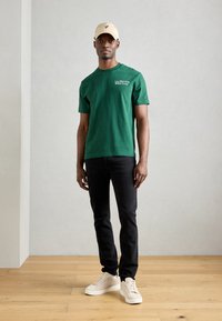 Grønn bomull t-skjorte med korte ermer og tekst på fronten, parret med svarte jeans og beige sneakers. Uformelt antrekk, minimalistisk design.