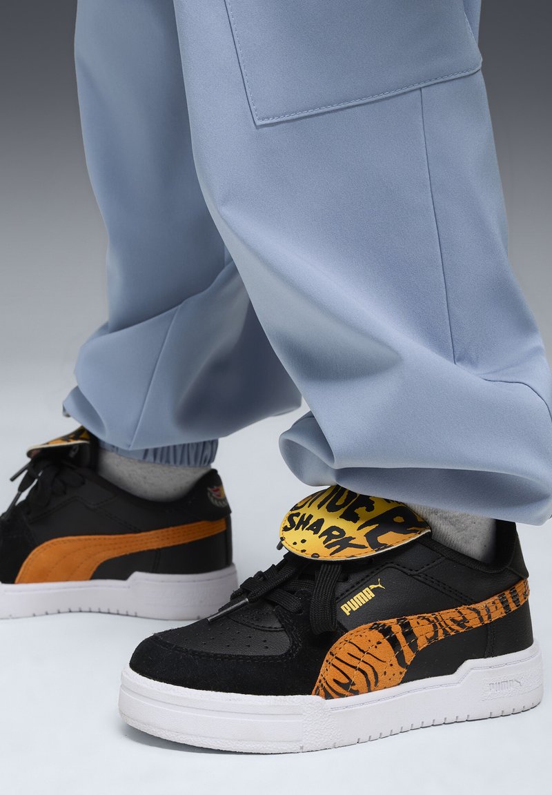 Baskets Puma noires et oranges avec des semelles blanches, motif rayé de tigre et texte "Shark" sur la languette, portées avec un pantalon bleu clair.